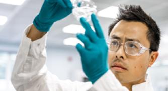 A Vertex scientist wearing a lab coat, goggles and gloves holds up a beaker with liquid in it